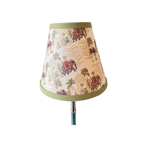 A conical table lamp shade with a cream-colored background and an all-over vintage-style print of red elephants, green palm trees, and figures. The shade is finished with olive green fabric trim at the top and bottom and is shown on a silver and teal lamp stem against a plain white background.