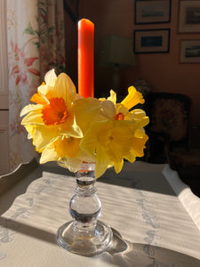 Yellow daffodils in a glass vase on a table with a candle and books in the background.